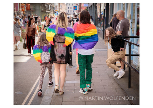 Three young women wearing rainbow wings walking away from a pride festival while a young girl looks on.
