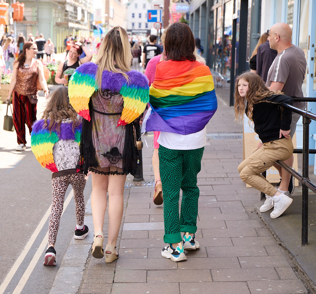 Three girls wearing rainbow angel wings walking down a street. 